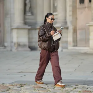 PARIS, FRANCE - DECEMBER 31: Yuwei Zhangzou wears hair pulled back into a low braided ponytail, makeup with an even complexion, defined brows and lipstick, wire earphones, oval dark-lens silver sunglasses by Celine, a brown oversized bomber leather jacket with a high collar and front zip closure, straight-leg brown sport jogger pants by Alo, a small structured beige and brown shearling fluffy bag with a top handle by Loro Piana, white shearling-lined lace-up shoes with brown suede panels, contrast laces and a flat rubber sole by Moon Boot, holds a mobile phone, during a street style fashion photo session, on December 31, 2025 in Paris, France. (Photo by Edward Berthelot/Getty Images)
