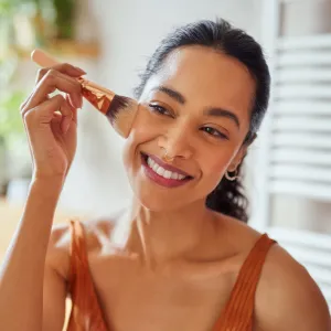 Beautiful girl applying makeup using powder brush before going to work. Healthy latin woman looking in the mirror and applying cosmetic with a big brush. Young woman looking in the mirror and applying foundation or blusher on her face.