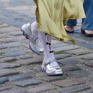 COPENHAGEN, DENMARK - AUGUST 06: Yamila Pavia wears a white blouse with voluminous sleeves and a high collar underneath a sleeveless, gathered dress in a mustard yellow shade. A large, sculptural crossbody bag in a gradient of purple hues is draped across the body. The hair is styled in a braided ponytail, adorned with small decorative elements. Footwear consists of metallic silver sneakers with a chunky sole, paired with white socks featuring a black pattern. A camera is held in one hand, and subtle makeup is applied, outside The Garment, during Copenhagen Fashion Week, on August 06, 2025 in Copenhagen, Denmark (Photo by Edward Berthelot/Getty Images)