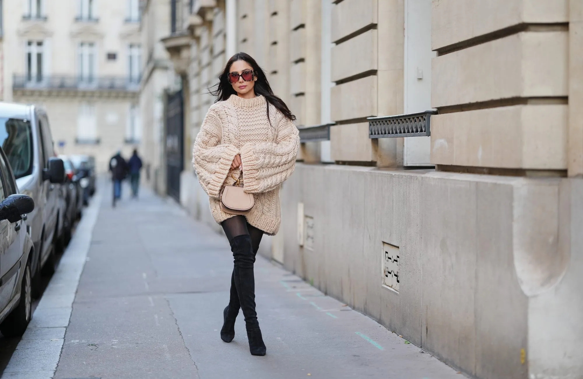 PARIS, FRANCE - DECEMBER 09: Patricia Gloria Contreras wears long straight dark brown hair worn loose, natural skin with neutral lipstick, oversized square burgundy tinted sunglasses, an oversized beige chunky knit sweater dress with a ribbed crew neckline, long ribbed cuffs and a textured cable knit pattern, sheer black tights, black suede over the knee boots, and a beige leather small top handle bag with a gold metal handle by Chloe, during a street style fashion photo session, on December 09, 2025 in Paris, France. (Photo by Edward Berthelot/Getty Images)