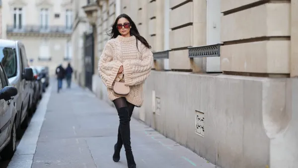 PARIS, FRANCE - DECEMBER 09: Patricia Gloria Contreras wears long straight dark brown hair worn loose, natural skin with neutral lipstick, oversized square burgundy tinted sunglasses, an oversized beige chunky knit sweater dress with a ribbed crew neckline, long ribbed cuffs and a textured cable knit pattern, sheer black tights, black suede over the knee boots, and a beige leather small top handle bag with a gold metal handle by Chloe, during a street style fashion photo session, on December 09, 2025 in Paris, France. (Photo by Edward Berthelot/Getty Images)