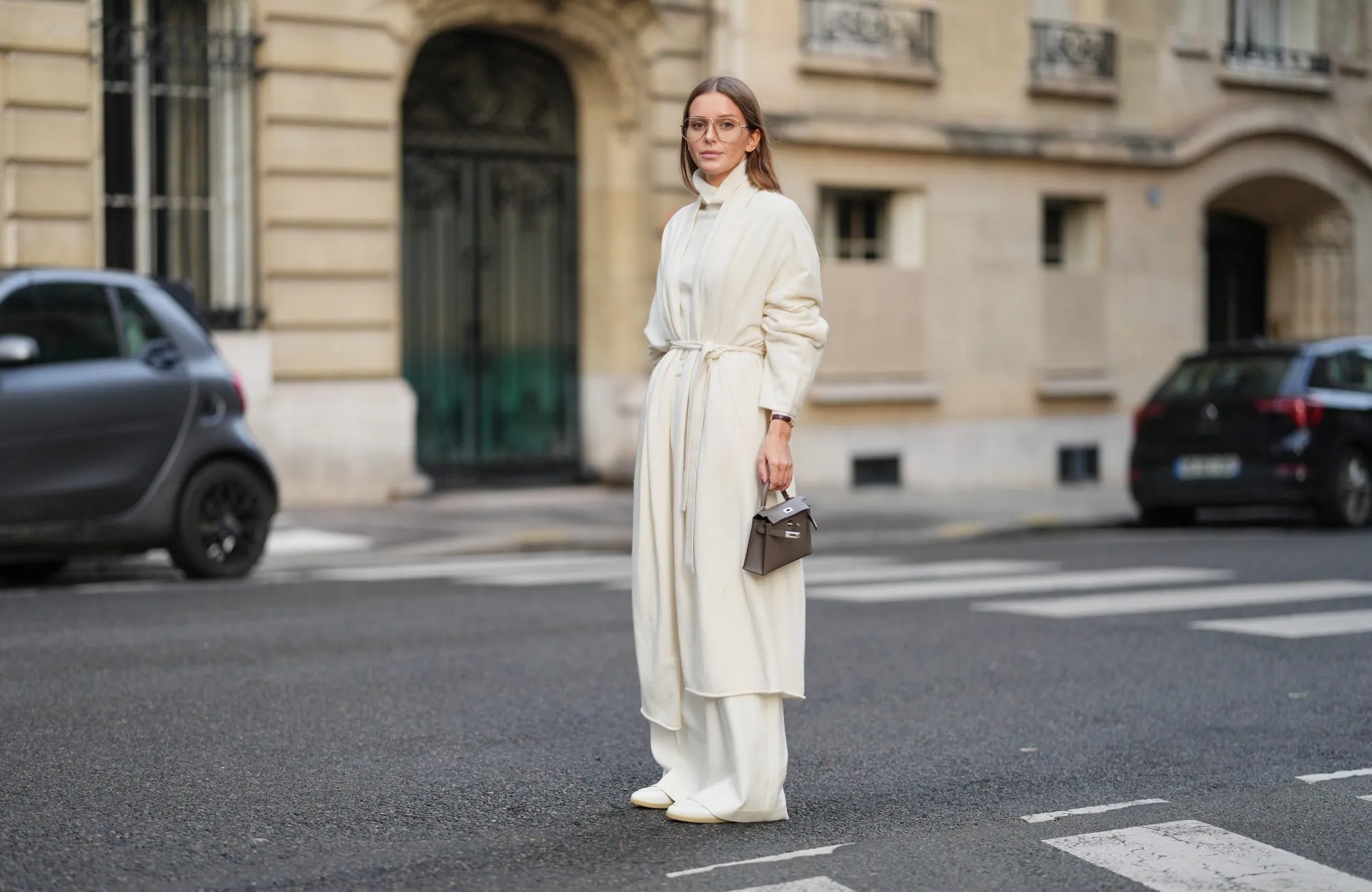 PARIS, FRANCE - DECEMBER 04: Diane Batoukina wears long light brown hair parted at the center with natural skin and neutral lipstick, a white cashmere long cardigan jacket by Lilysilk, a white cashmere turtleneck sweater by Bompard, white straight pants by Lilysilk, white leather loafers shoes by Baudoin and Lange, a grey Hermes Kelly 20 bag, during a street style fashion photo session, on December 04, 2025 in Paris, France, full body shot. (Photo by Edward Berthelot/Getty Images)