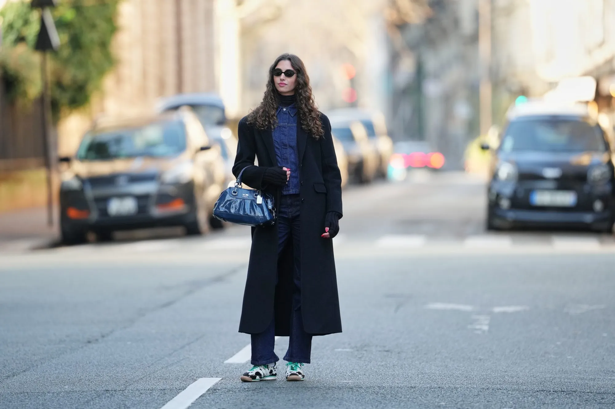 PARIS, FRANCE - DECEMBER 27: Amanda Derhy wears long curly / wavy brown hair worn loose with a center part, black oval sunglasses from Miu Miu, make-up is a natural complexion with defined brows and soft pink lipstick, a black wool winter long coat, a black ribbed turtleneck top layered under a dark indigo blue denim jacket with a pointed collar, vertical seam detailing and a fitted silhouette, a dark blue leather Coach bag with metal ring handles and visible logo plaque, matching straight-leg dark indigo denim jeans pants, Adidas SL72 OG sneakers shoes in white with green shoelaces and contrasting black detailing / cow pattern print, during a street style fashion photo session, on December 27, 2025 in Paris, France. (Photo by Edward Berthelot/Getty Images)