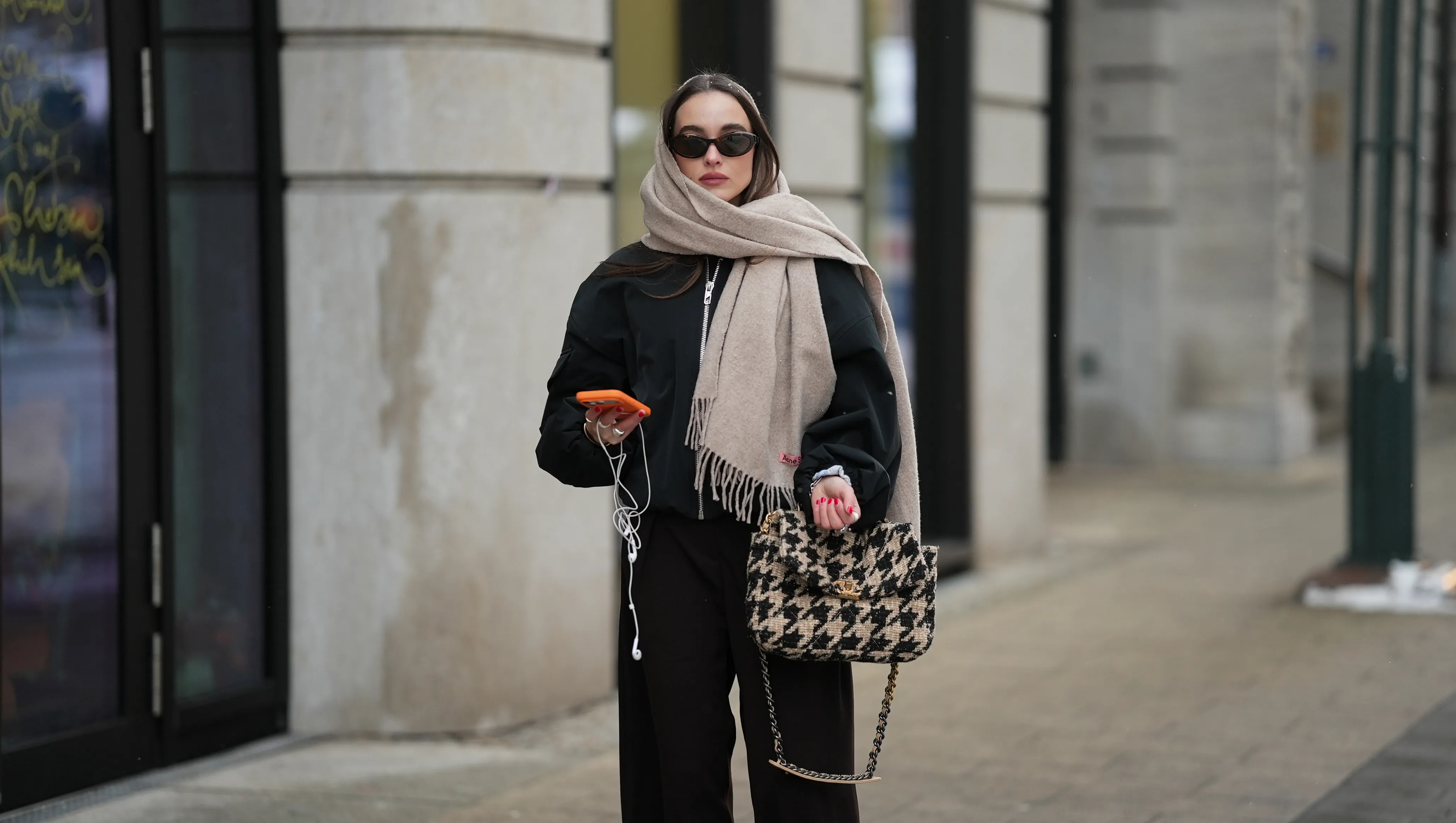 HAMBURG, GERMANY - JANUARY 5: Evi Wave is seen wearing a black bomber jacket with a relaxed oversized silhouette, dropped shoulders, elasticated cuffs and a zip front closure from GANNI; a beige wool scarf with a soft texture, long fringed edges wrapped around the neck and head from Acne Studios; dark brown wide leg trousers with a fluid drape and straight loose cut from Anine Bing; a black and beige houndstooth patterned handbag with a structured rectangular shape, woven textile surface, gold metal chain shoulder strap and gold CC logo frontal clasp called ‚ÄúChanel 19 Medium‚Äù from Chanel; an iPhone 17 Pro with a bright orange phone case from Apple; white wired headphones from Apple; among her jewelry are chunky silver rings; oval and narrow sunglasses with a dark tortoiseshell patterned acetate frame and dark lenses from Prada; her long dark brown straight hair is worn loose and parted in the middle covered by the scarf on January 5, 2026 in Hamburg, Germany. (Photo by Moritz Scholz/Getty Images)