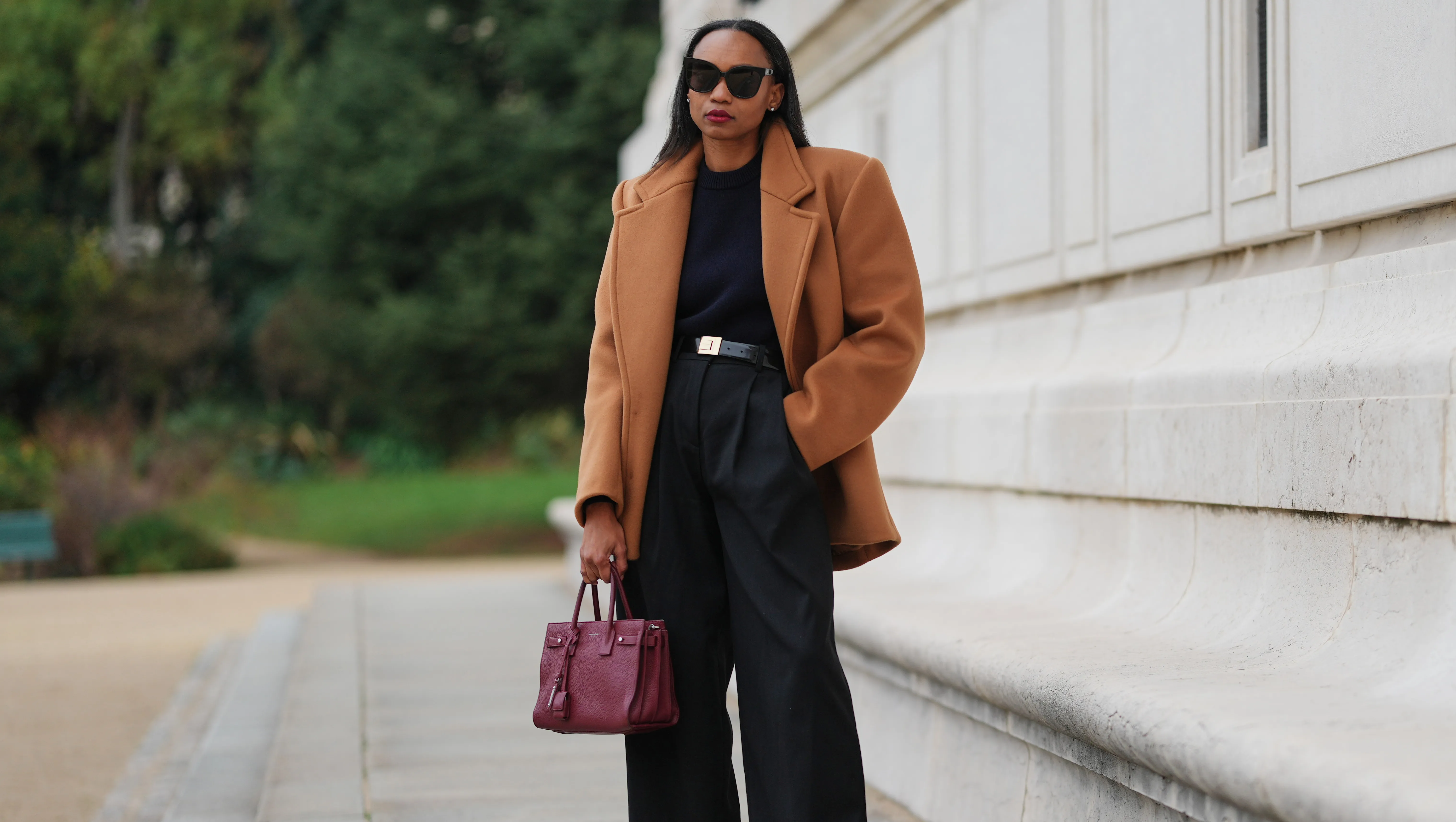 PARIS, FRANCE - DECEMBER 13: Emilie Joseph wears long straight dark brown hair parted at the center, makeup featuring an even complexion with matte skin and deep red lipstick, oversized black sunglasses by Gentle Monster, a camel sculptural wool coat by Saint Laurent with sharp tailoring and a martingale detail, a chocolate brown midi skirt with a fringe hem, a structured navy wool sweater with shoulder pads by COS, high-waisted black wool trousers by COS, a polished black leather belt with an oversized engraved gold buckle by Saint Laurent, a burgundy leather handbag by YSL Saint Laurent, and burgundy patent leather Blake slingback heels by Saint Laurent, during a street style fashion photo session on December 13, 2025 in Paris, France. (Photo by Edward Berthelot/Getty Images)