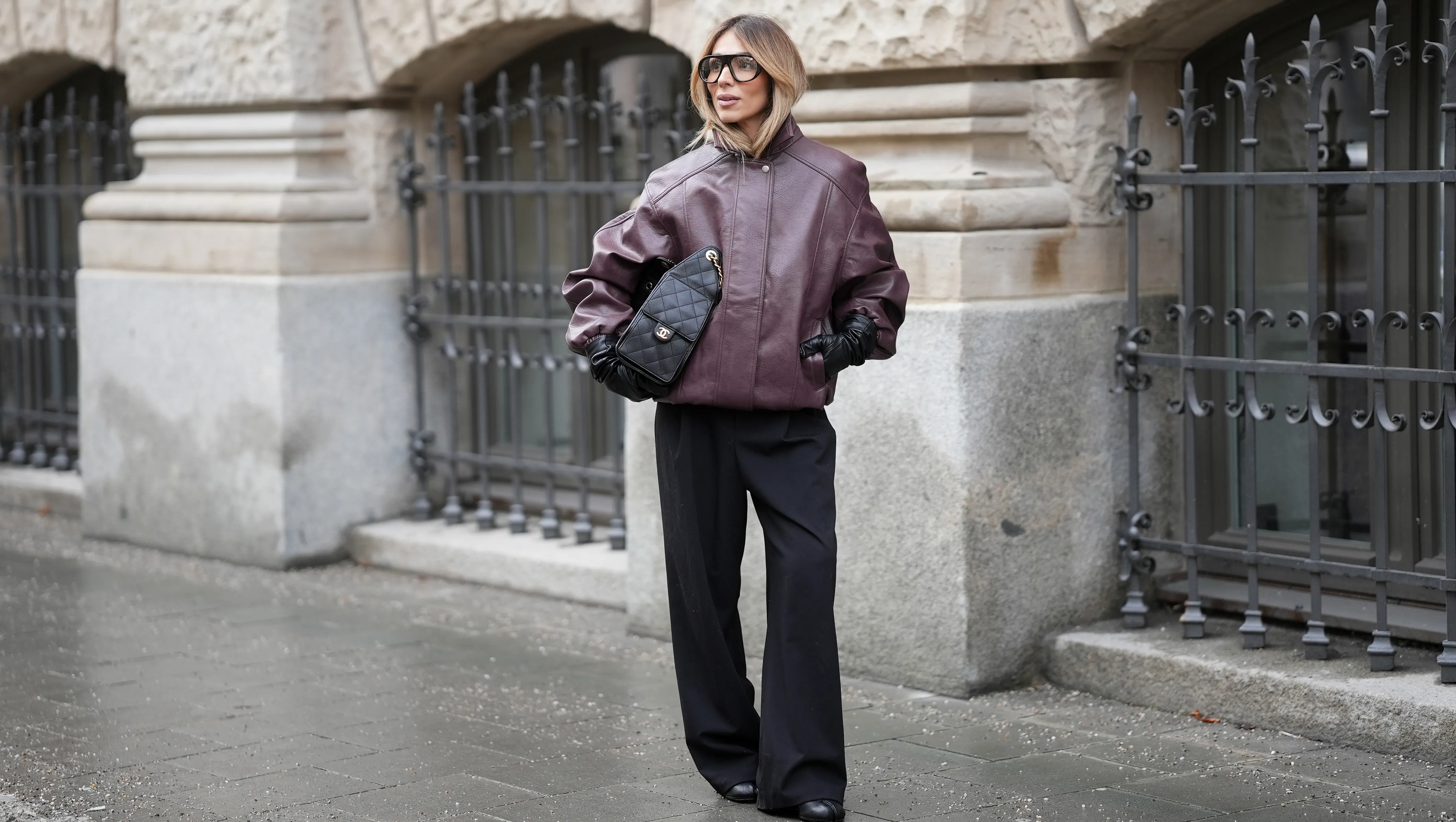 MUNICH, GERMANY - JANUARY 10: Oeznur Sinani alias Sinperi is seen wearing an oversized burgundy faux leather Prescott biker jacket with collar, hidden button front closure and minimalist design from The Frankie Shop; wide-leg black tailored trousers with relaxed fit from COS; black leather Tabi boots with flat sole and split toe from Maison Margiela; a black quilted grained calfskin leather Chanel 25 medium handbag with flap pockets, drawstring closing and golden chain strap and logo detail from Chanel; long black leather gloves from Mango; black oversized aviator glasses with black acetate frames and clear glasses from Prada; her blonde hair is styled in a blow out look on January 10, 2026 in Munich, Germany. (Photo by Moritz Scholz/Getty Images)