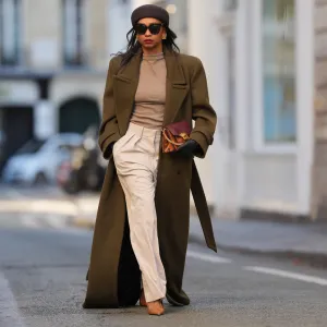 PARIS, FRANCE - JANUARY 03: Emilie Joseph wears straight black hair worn loose under a brown wool pillbox hat by COS, black rectangular sunglasses, a natural makeup look with defined brows, even skin, deep red lipstick, gold-tone statement earrings, an oversized military-style wool long coat in deep olive green with sharp structured shoulders by Saint Laurent, a fitted taupe mock-neck knit top, high-waisted wide-leg cream white canvas trousers with front pleats by The Frankie Shop, a burgundy and python-embossed leather clutch bag with a gold-tone buckle by Givenchy, black leather gloves, pointed-toe leather slingback heels shoes in cognac brown by Saint Laurent, during a street style photo session, on January 03, 2026 in Paris, France.