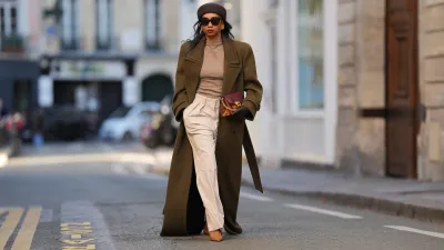 PARIS, FRANCE - JANUARY 03: Emilie Joseph wears straight black hair worn loose under a brown wool pillbox hat by COS, black rectangular sunglasses, a natural makeup look with defined brows, even skin, deep red lipstick, gold-tone statement earrings, an oversized military-style wool long coat in deep olive green with sharp structured shoulders by Saint Laurent, a fitted taupe mock-neck knit top, high-waisted wide-leg cream white canvas trousers with front pleats by The Frankie Shop, a burgundy and python-embossed leather clutch bag with a gold-tone buckle by Givenchy, black leather gloves, pointed-toe leather slingback heels shoes in cognac brown by Saint Laurent, during a street style photo session, on January 03, 2026 in Paris, France.