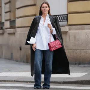 PARIS, FRANCE – DECEMBER 18: Diane Batoukina wears straight brown hair worn loose and parted at the center, clear eyeglasses, a white cotton button-up shirt, a long black leather coat, dark blue wide-leg denim jeans, black pointed-toe leather shoes, a red leather Chanel bag, during a street style fashion photo session, on December 18, 2025 in Paris, France.