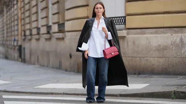PARIS, FRANCE – DECEMBER 18: Diane Batoukina wears straight brown hair worn loose and parted at the center, clear eyeglasses, a white cotton button-up shirt, a long black leather coat, dark blue wide-leg denim jeans, black pointed-toe leather shoes, a red leather Chanel bag, during a street style fashion photo session, on December 18, 2025 in Paris, France.