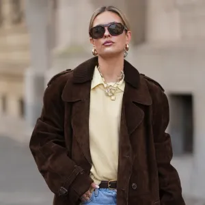 FRANKFURT AM MAIN, GERMANY - JANUARY 21: Julia Kammerer is seen wearing a long chocolate brown suede vintage trench coat with a wide lapel collar, button front and a relaxed fit; a butter yellow blouse with classic collar from JJXX; a light yellow knit sweater from H&M layered above the blouse; light blue wash jeans with turned up cuffs, straight leg and relaxed fit from Levi’s; a mocha brown suede leather Midi Hudson tote bag with top handle and adjustable belt from DeMellier; oversized aviator sunglasses with brown tortoiseshell frames and tinted lenses from H&M; a gold-plated brass necklace with natural freshwater pearls from Scandivv; a golden chain necklace with key lock pendant from MyJewellery; a golden chain necklace with „A“-letter pendant from Scandivv; chunky golden hoop earrings from COS; a slim brown leather belt with golden buckle from Lilysilk; Samba OG leather-trimmed leopard-print pony hair sneakers from Adidas x Wales Bonner; her blonde hair styled back on January 21, 2026 in Frankfurt am Main, Germany. (Photo by Moritz Scholz/Getty Images)