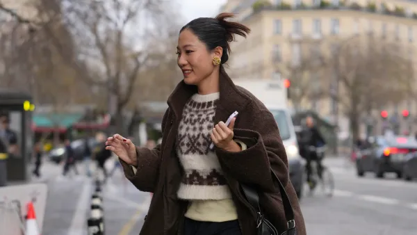PARIS, FRANCE - JANUARY 23: A guest wears dark brown hair pulled back into a mid ponytail, gold metal statement earrings, a black leather shoulder bag, a brown wool coat, a white knit sweater with a brown patterned motif and a pale yellow knit top, dark blue pinstriped trousers, black leather knee high boots shoes, outside Maison Mihara Yasuhiro, during Paris Fashion Week - Menswear Fall/Winter 2026-2027, on January 23, 2026 in Paris, France (Photo by Edward Berthelot/Getty Images)