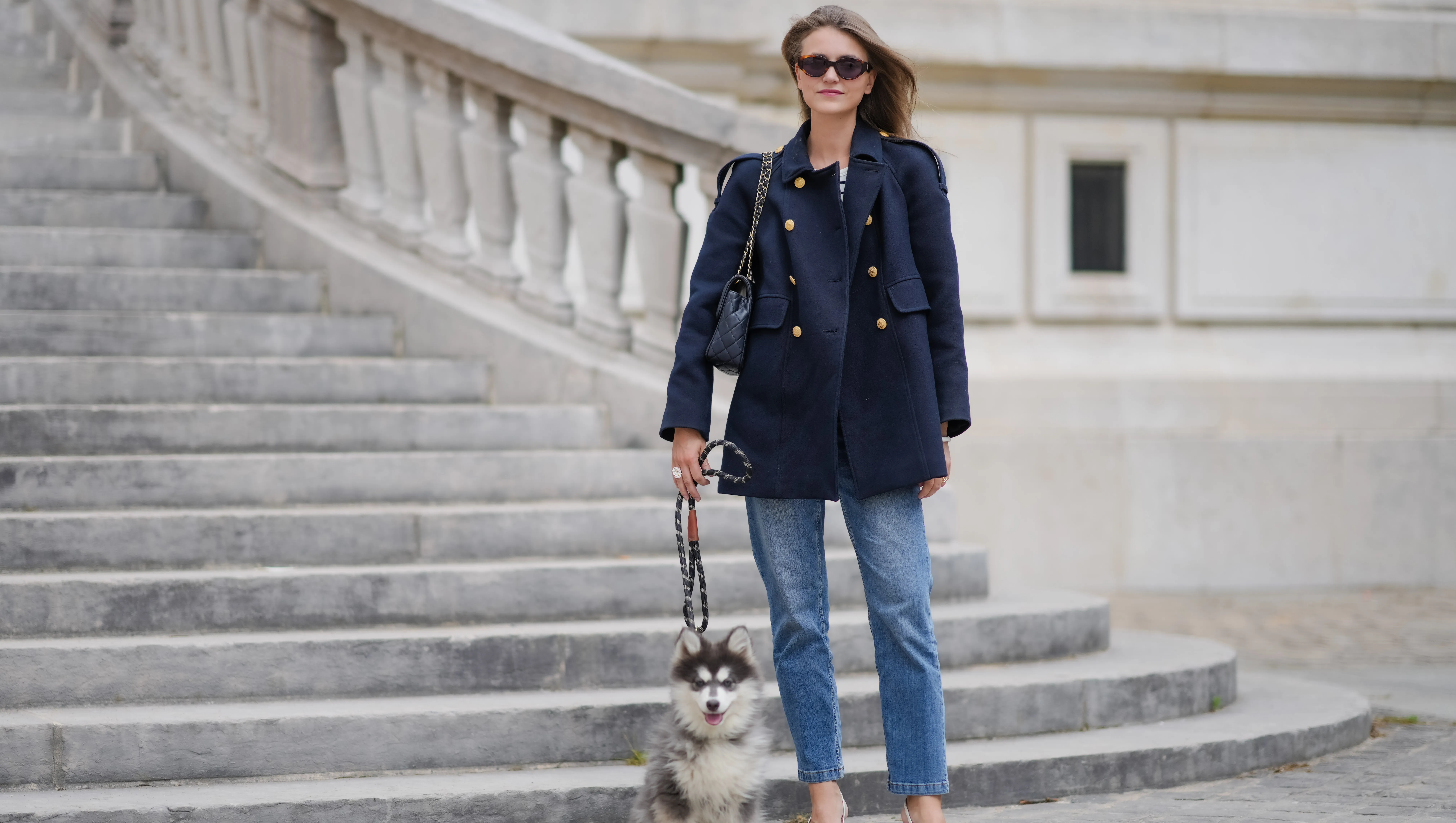 PARIS, FRANCE - MAY 20: Segolene Hyppolite wears sunglasses, a dark navy blue pea coat / peacoat jacket with golden buttons from Tara Jarmon, a black and white top with stripes from Sezane, blue denim pants / jeans from Vanessa Bruno, pointed shoes / pumps from Jonak, a Chanel bag, and holds her husky / dog /pet with a leash, during a street style fashion photo session, on May 20, 2024 in Paris, France. (Photo by Edward Berthelot/Getty Images)