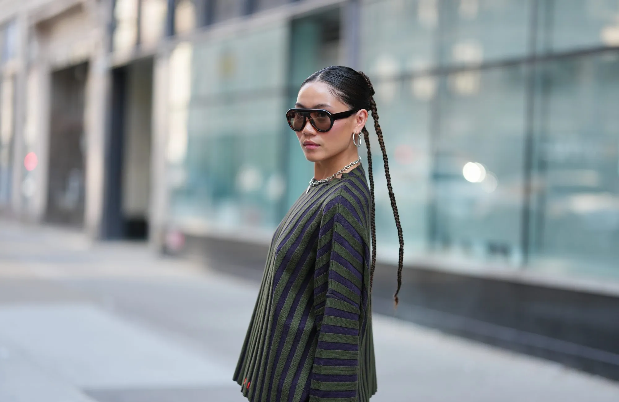 NEW YORK, NEW YORK - SEPTEMBER 14: A guest wears a center-parted slick low bun, oversized dark brown gradient sunglasses, a chunky silver chain necklace with a small round silver pendant and medium silver hoop earrings, a ribbed knit long-sleeve sweater with vertical dark navy and olive green stripes over olive green shorts, outside Gabe Gordon, during New York Fashion Week, on September 14, 2025 in New York, New York (Photo by Edward Berthelot/Getty Images)