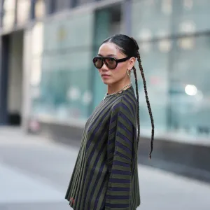 NEW YORK, NEW YORK - SEPTEMBER 14: A guest wears a center-parted slick low bun, oversized dark brown gradient sunglasses, a chunky silver chain necklace with a small round silver pendant and medium silver hoop earrings, a ribbed knit long-sleeve sweater with vertical dark navy and olive green stripes over olive green shorts, outside Gabe Gordon, during New York Fashion Week, on September 14, 2025 in New York, New York (Photo by Edward Berthelot/Getty Images)