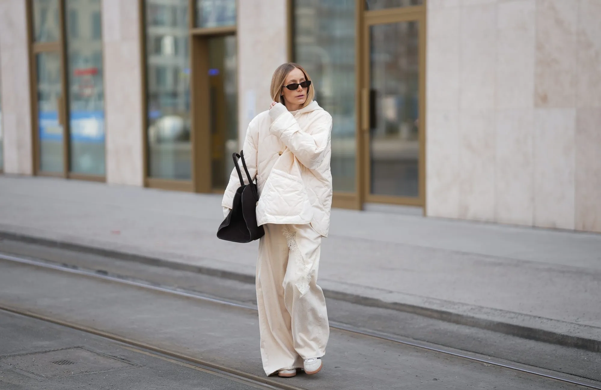 MUNICH, GERMANY - JANUARY 22: Anouk Delphine is seen wearing a thin cloud dancer white quilted puffer jacket with button front and oversized fit from The Frankie Shop; a cream white lace-trimmed top with asymmetric hemline from Zara; a cream knit sweater with hood from Windsor; off-white wide-leg trousers with vertical seam details and flared hems from H&M; slim sunglasses with black acetate frame and dark tinted lenses from Gucci; a black suede leather tote bag with top handles and slightly trapezoidal from COS; white Samba OG suede sneakers with black stripes from Adidas; her blonde hair is worn straight and parted in the middle on January 22, 2026 in Munich, Germany. (Photo by Moritz Scholz/Getty Images)