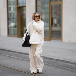 MUNICH, GERMANY - JANUARY 22: Anouk Delphine is seen wearing a thin cloud dancer white quilted puffer jacket with button front and oversized fit from The Frankie Shop; a cream white lace-trimmed top with asymmetric hemline from Zara; a cream knit sweater with hood from Windsor; off-white wide-leg trousers with vertical seam details and flared hems from H&M; slim sunglasses with black acetate frame and dark tinted lenses from Gucci; a black suede leather tote bag with top handles and slightly trapezoidal from COS; white Samba OG suede sneakers with black stripes from Adidas; her blonde hair is worn straight and parted in the middle on January 22, 2026 in Munich, Germany. (Photo by Moritz Scholz/Getty Images)
