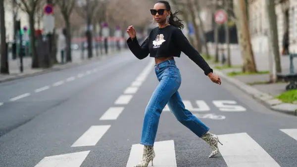 Emilie Joseph wears sunglasses, Isabel Marant bejeweled long earrings, a cropped black pullover with printed angels from Fiorucci, blue ribcage Levi's denim jeans, white high heels pointy ankle boots with snake print from Paris Texas, on February 16, 2021 in Paris, France.