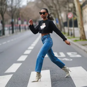Emilie Joseph wears sunglasses, Isabel Marant bejeweled long earrings, a cropped black pullover with printed angels from Fiorucci, blue ribcage Levi's denim jeans, white high heels pointy ankle boots with snake print from Paris Texas, on February 16, 2021 in Paris, France.