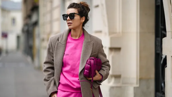 Katie Giorgadze @katie.one wears a black sunglasses, a neon pink recycled cotton sweater, matching neon pink recycled cotton sport pants / jogger pants, a pale brown wool long coat, a purple shiny leather Padded Cassette shoulder bag from Bottega Veneta, during a street style fashion photo session, on December 11, 2021 in Paris, France.