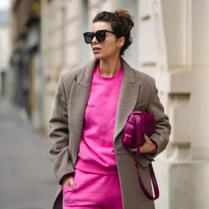 Katie Giorgadze @katie.one wears a black sunglasses, a neon pink recycled cotton sweater, matching neon pink recycled cotton sport pants / jogger pants, a pale brown wool long coat, a purple shiny leather Padded Cassette shoulder bag from Bottega Veneta, during a street style fashion photo session, on December 11, 2021 in Paris, France.
