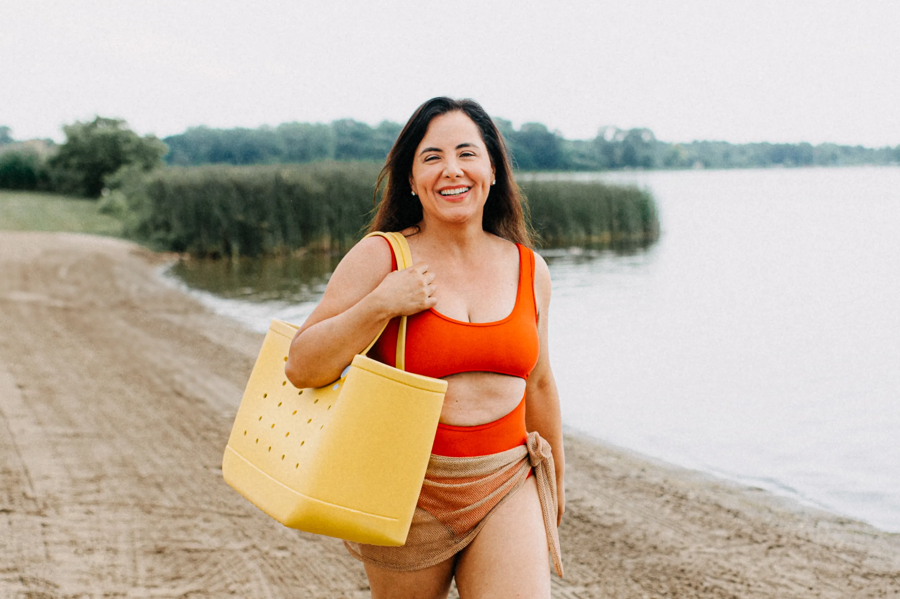 A woman in a red swimsuit carries a large yellow tote bag while walking along a sandy beach with greenery and water in the background. She appears to be enjoying a day outdoors, exhibiting a cheerful expression.