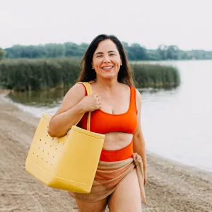 A woman in a red swimsuit carries a large yellow tote bag while walking along a sandy beach with greenery and water in the background. She appears to be enjoying a day outdoors, exhibiting a cheerful expression.