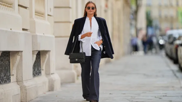 Diane Batoukina wears light brown hair styled straight with a middle part, dark rectangular sunglasses by Montblanc, a black blazer jacket by Anine Bing draped over the shoulders, a white pleated blouse by Bourrienne Paris, dark blue wide-leg jeans by APC, a black quilted leather Chanel bag with chain strap, and brown pointed-toe heels by Manolo Blahnik, during a street style fashion photo session, on September 05, 2025 in Paris, France.
