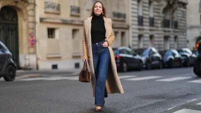 Alba Garavito Torre wears long brown hair parted at the center with natural skin and neutral lipstick, a black long-sleeve turtleneck pullover by Citizens of Humanity, dark-indigo high-rise flare jeans pants by Citizens of Humanity, a beige long shearling coat jacket by Possery, brown patent-leather shoes by Jonak, a brown suede New York midi bag by Demellier London, gold pearl earrings and gold rings by Sempiterno Paris, during a street style fashion photo session, on December 04, 2025 in Paris, France, full body shot.