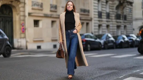 Alba Garavito Torre wears long brown hair parted at the center with natural skin and neutral lipstick, a black long-sleeve turtleneck pullover by Citizens of Humanity, dark-indigo high-rise flare jeans pants by Citizens of Humanity, a beige long shearling coat jacket by Possery, brown patent-leather shoes by Jonak, a brown suede New York midi bag by Demellier London, gold pearl earrings and gold rings by Sempiterno Paris, during a street style fashion photo session, on December 04, 2025 in Paris, France, full body shot.