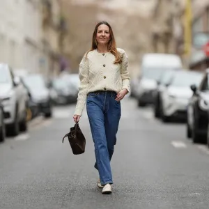 Alba Garavito Torre wears long light brown hair worn loose and parted at the side, small gold-tone earrings by Sempiterno Paris, an ecru white woven cable-knit cardigan with a round neckline, textured panels and front button fastening, dark blue straight-leg denim 501 jeans by Levis with a classic five-pocket construction, ecru patent leather loafers by Jonak with a rounded toe and low heel, a chocolate brown suede bucket bag by Saonara with a drawstring closure and top handle carried by hand, during a street style fashion photo session, on January 15, 2026 in Paris, France.