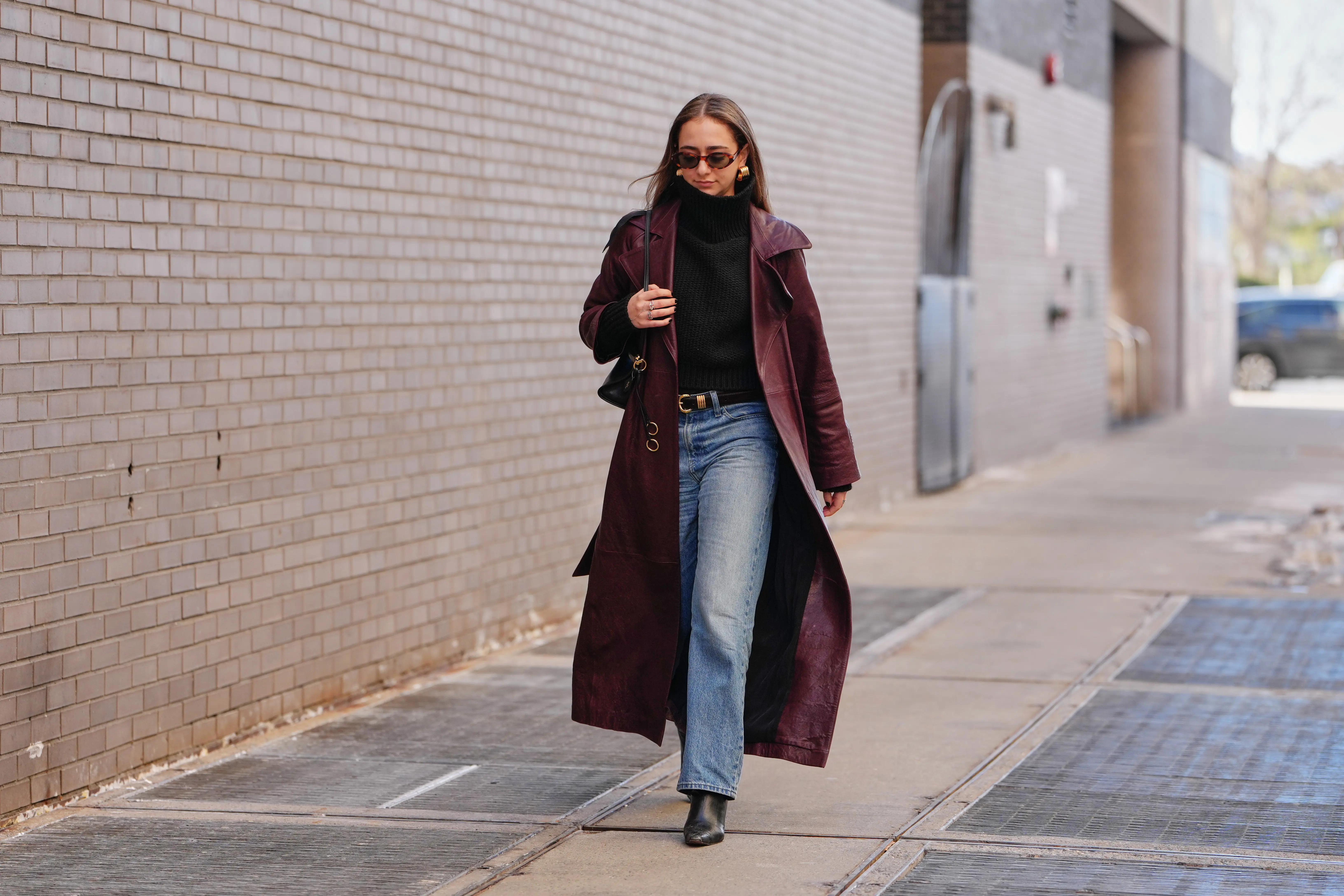 A guest wears long hair, brown tinted sunglasses, earrings, rings, a black leather bag, a red burgundy leather coat, a black knit high neck sweater, blue light wash straight-leg jeans pants, a black shiny leather belt, black leather pointed-toe ankle boots shoes, outside 7 for All Mankind, during New York Fashion Week, on February 13, 2026 in New York City