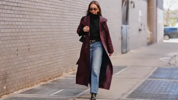 A guest wears long hair, brown tinted sunglasses, earrings, rings, a black leather bag, a red burgundy leather coat, a black knit high neck sweater, blue light wash straight-leg jeans pants, a black shiny leather belt, black leather pointed-toe ankle boots shoes, outside 7 for All Mankind, during New York Fashion Week, on February 13, 2026 in New York City