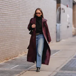 A guest wears long hair, brown tinted sunglasses, earrings, rings, a black leather bag, a red burgundy leather coat, a black knit high neck sweater, blue light wash straight-leg jeans pants, a black shiny leather belt, black leather pointed-toe ankle boots shoes, outside 7 for All Mankind, during New York Fashion Week, on February 13, 2026 in New York City