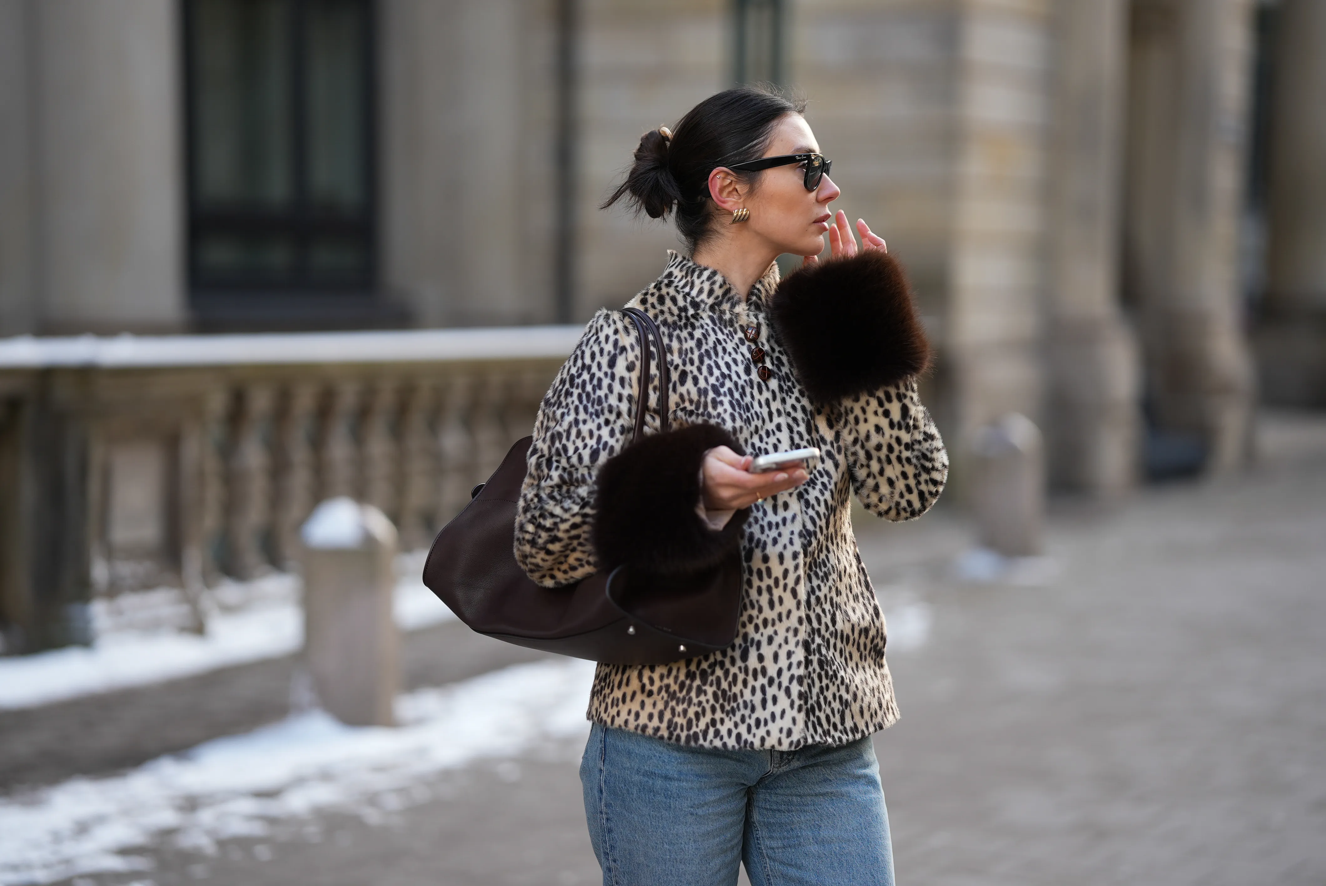 Milena Karl is seen wearing a cream and black leopard print "dolly" faux fur jacket with a classic collar, front leather button closure and dark brown fur trimmed cuffs from Carelli; light blue straight leg jeans with a mid rise fit from Massimo Dutti; the &ldquo;Soft Margaux 15&rdquo; handbag crafted in smooth dark brown leather with a softly structured silhouette, side buckle detailing, curved top handles and subtle logo detailing from The Row; holding an iPhone from Apple; rectangular black "Wayfarer" sunglasses with dark tinted lenses from Ray-Ban; sculptural gold colored earrings from Mango; her dark brown hair styled in a sleek low bun on February 14, 2026 in Hamburg, Germany.