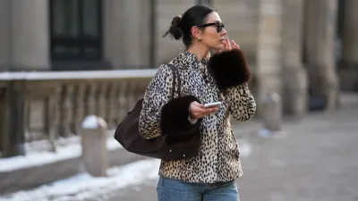 Milena Karl is seen wearing a cream and black leopard print "dolly" faux fur jacket with a classic collar, front leather button closure and dark brown fur trimmed cuffs from Carelli; light blue straight leg jeans with a mid rise fit from Massimo Dutti; the “Soft Margaux 15” handbag crafted in smooth dark brown leather with a softly structured silhouette, side buckle detailing, curved top handles and subtle logo detailing from The Row; holding an iPhone from Apple; rectangular black "Wayfarer" sunglasses with dark tinted lenses from Ray-Ban; sculptural gold colored earrings from Mango; her dark brown hair styled in a sleek low bun on February 14, 2026 in Hamburg, Germany.
