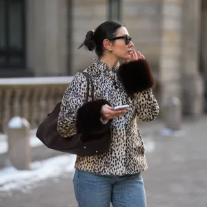 Milena Karl is seen wearing a cream and black leopard print "dolly" faux fur jacket with a classic collar, front leather button closure and dark brown fur trimmed cuffs from Carelli; light blue straight leg jeans with a mid rise fit from Massimo Dutti; the “Soft Margaux 15” handbag crafted in smooth dark brown leather with a softly structured silhouette, side buckle detailing, curved top handles and subtle logo detailing from The Row; holding an iPhone from Apple; rectangular black "Wayfarer" sunglasses with dark tinted lenses from Ray-Ban; sculptural gold colored earrings from Mango; her dark brown hair styled in a sleek low bun on February 14, 2026 in Hamburg, Germany.