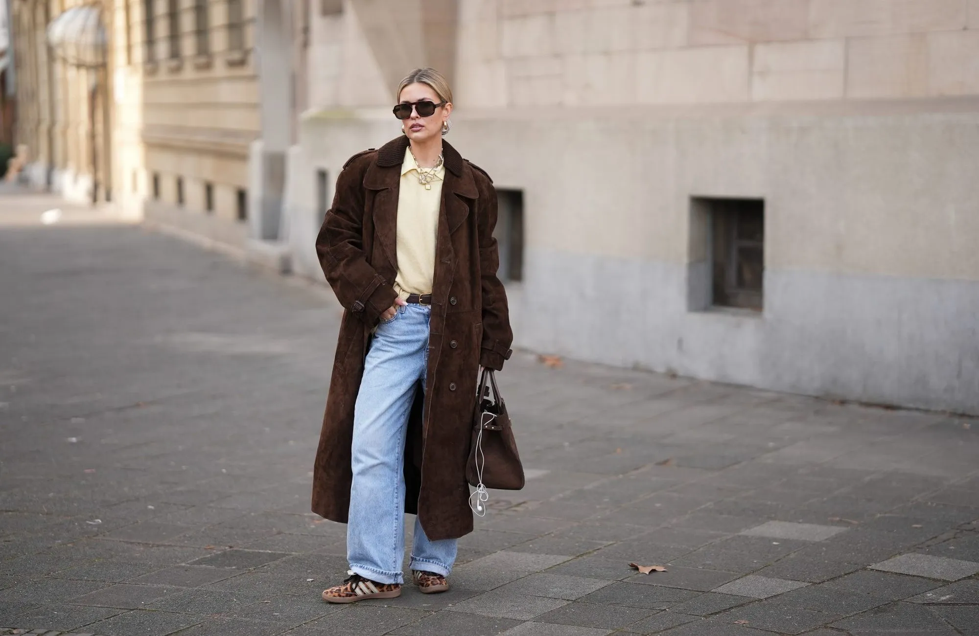 FRANKFURT AM MAIN, GERMANY - JANUARY 21: Julia Kammerer is seen wearing a long chocolate brown suede vintage trench coat with a wide lapel collar, button front and a relaxed fit; a butter yellow blouse with classic collar from JJXX; a light yellow knit sweater from H&M layered above the blouse; light blue wash jeans with turned up cuffs, straight leg and relaxed fit from Levi’s; a mocha brown suede leather Midi Hudson tote bag with top handle and adjustable belt from DeMellier; oversized aviator sunglasses with brown tortoiseshell frames and tinted lenses from H&M; a gold-plated brass necklace with natural freshwater pearls from Scandivv; a golden chain necklace with key lock pendant from MyJewellery; a golden chain necklace with „A“-letter pendant from Scandivv; chunky golden hoop earrings from COS; a slim brown leather belt with golden buckle from Lilysilk; Samba OG leather-trimmed leopard-print pony hair sneakers from Adidas x Wales Bonner; her blonde hair styled back on January 21, 2026 in Frankfurt am Main, Germany. (Photo by Moritz Scholz/Getty Images)