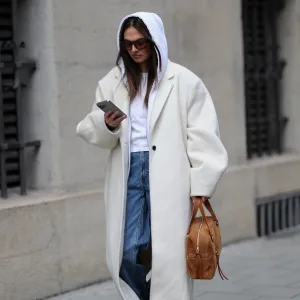 MUNICH, GERMANY - FEBRUARY 3: Anna Winter is seen wearing a long ivory colored wool coat called "NUEVO COCOON COAT, POWDER" with dropped padded shoulders, wide lapels, single button closure and an oversized straight silhouette from Source Unknown; a white hooded zip jacket in a cotton fabric from Brandy Melville; a white T-shirt from Brandy Melville; mid blue straight leg denim jeans with a relaxed fit from Weekday; cream colored leather sneakers with a rubber sole from New Balance; the "Lucky Bowling shoulder bag" in brown suede leather with gold hardware and front zip pocket detailing from Jérôme Dreyfuss; holding an iPhone from Apple; square acetate sunglasses with brown tinted lenses from Vehla; her brown hair is worn loose and straight with a center part covered by the hood on February 3, 2026 in Munich, Germany. (Photo by Moritz Scholz/Getty Images)