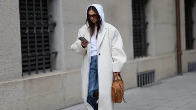 MUNICH, GERMANY - FEBRUARY 3: Anna Winter is seen wearing a long ivory colored wool coat called "NUEVO COCOON COAT, POWDER" with dropped padded shoulders, wide lapels, single button closure and an oversized straight silhouette from Source Unknown; a white hooded zip jacket in a cotton fabric from Brandy Melville; a white T-shirt from Brandy Melville; mid blue straight leg denim jeans with a relaxed fit from Weekday; cream colored leather sneakers with a rubber sole from New Balance; the "Lucky Bowling shoulder bag" in brown suede leather with gold hardware and front zip pocket detailing from Jérôme Dreyfuss; holding an iPhone from Apple; square acetate sunglasses with brown tinted lenses from Vehla; her brown hair is worn loose and straight with a center part covered by the hood on February 3, 2026 in Munich, Germany. (Photo by Moritz Scholz/Getty Images)