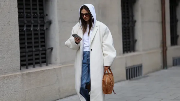 MUNICH, GERMANY - FEBRUARY 3: Anna Winter is seen wearing a long ivory colored wool coat called "NUEVO COCOON COAT, POWDER" with dropped padded shoulders, wide lapels, single button closure and an oversized straight silhouette from Source Unknown; a white hooded zip jacket in a cotton fabric from Brandy Melville; a white T-shirt from Brandy Melville; mid blue straight leg denim jeans with a relaxed fit from Weekday; cream colored leather sneakers with a rubber sole from New Balance; the "Lucky Bowling shoulder bag" in brown suede leather with gold hardware and front zip pocket detailing from Jérôme Dreyfuss; holding an iPhone from Apple; square acetate sunglasses with brown tinted lenses from Vehla; her brown hair is worn loose and straight with a center part covered by the hood on February 3, 2026 in Munich, Germany. (Photo by Moritz Scholz/Getty Images)