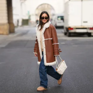 BERLIN, GERMANY - NOVEMBER 25: Laura Noltemeyer is seen wearing a shearling lining coat in a rich cognac tone with an oversized straight silhouette, wide lapels and buckle-strap cuff detailing on the sleeves from Max&Co; layered underneath is a soft beige wool-blend knit sweater with a ribbed crew neckline and ribbed trims from Sézane; dark-blue straight-leg denim trousers with a classic fit from By Aylin Koenig; beige suede winter boots with a rounded toe, exposed stitching and shearling lining from Inuikii; a white structured leather top-handle bag in an intrecciato weave and trapeze silhouette called “Intrecciato Tote” from Bottega Veneta; chunky sculptural gold earrings from Zara; black angular acetate sunglasses with a thick frame and dark lenses from Saint Laurent; her dark-brown hair is worn straight with a center part on November 25, 2025 in Berlin, Germany. (Photo by Moritz Scholz/Getty Images)