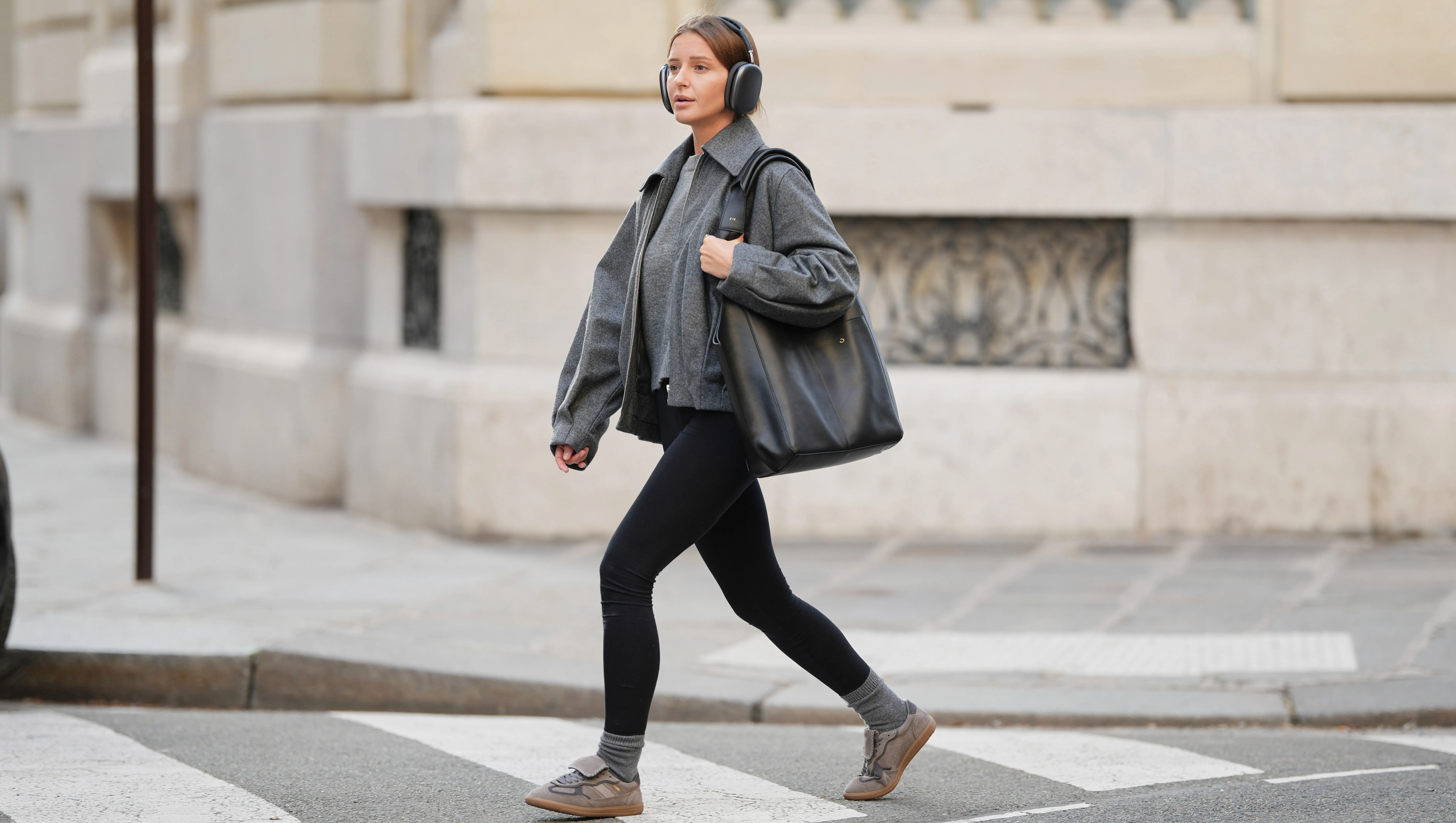 PARIS, FRANCE - JANUARY 15: Diane Batoukina wears long straight brown hair pulled back with a center part, over-ear silver headphones Apple airpod Max, a medium grey knit sweater by Zara with a crew neckline layered under a dark grey wool zip-front jacket by House of Dagmar featuring a pointed collar and relaxed fit, black stretch leggings by Zara with a matte finish, ribbed grey cashmere socks by Mango, grey and taupe lace-up sneakers by Alohas with a low-profile rubber sole, and a large black leather RSVP tote bag with long shoulder straps carried at the side, during a street style fashion photo session, on January 15, 2026 in Paris, France. (Photo by Edward Berthelot/Getty Images)