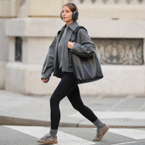 PARIS, FRANCE - JANUARY 15: Diane Batoukina wears long straight brown hair pulled back with a center part, over-ear silver headphones Apple airpod Max, a medium grey knit sweater by Zara with a crew neckline layered under a dark grey wool zip-front jacket by House of Dagmar featuring a pointed collar and relaxed fit, black stretch leggings by Zara with a matte finish, ribbed grey cashmere socks by Mango, grey and taupe lace-up sneakers by Alohas with a low-profile rubber sole, and a large black leather RSVP tote bag with long shoulder straps carried at the side, during a street style fashion photo session, on January 15, 2026 in Paris, France. (Photo by Edward Berthelot/Getty Images)