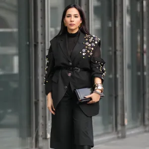PARIS, FRANCE - JANUARY 20: Samia Laaboudi wears long straight dark brown hair with a center part, metal hoop earrings, a black leather clutch bag with gold tone hardware, a black sheer mesh turtleneck top under a black tailored belted blazer jacket with notch lapels and rolled sleeves embellished with gold tone studs and round metallic badges, black wide leg culotte knee length cropped trousers, black leather knee high boots shoes with a pointed toe and block heel, outside Etudes, during Paris Fashion Week - Menswear Fall/Winter 2026-2027, on January 20, 2026 in Paris, France (Photo by Edward Berthelot/Getty Images)