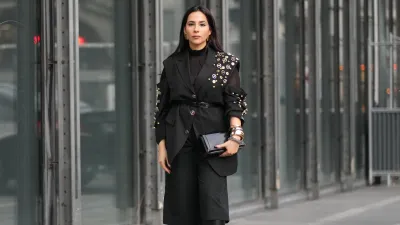 PARIS, FRANCE - JANUARY 20: Samia Laaboudi wears long straight dark brown hair with a center part, metal hoop earrings, a black leather clutch bag with gold tone hardware, a black sheer mesh turtleneck top under a black tailored belted blazer jacket with notch lapels and rolled sleeves embellished with gold tone studs and round metallic badges, black wide leg culotte knee length cropped trousers, black leather knee high boots shoes with a pointed toe and block heel, outside Etudes, during Paris Fashion Week - Menswear Fall/Winter 2026-2027, on January 20, 2026 in Paris, France (Photo by Edward Berthelot/Getty Images)