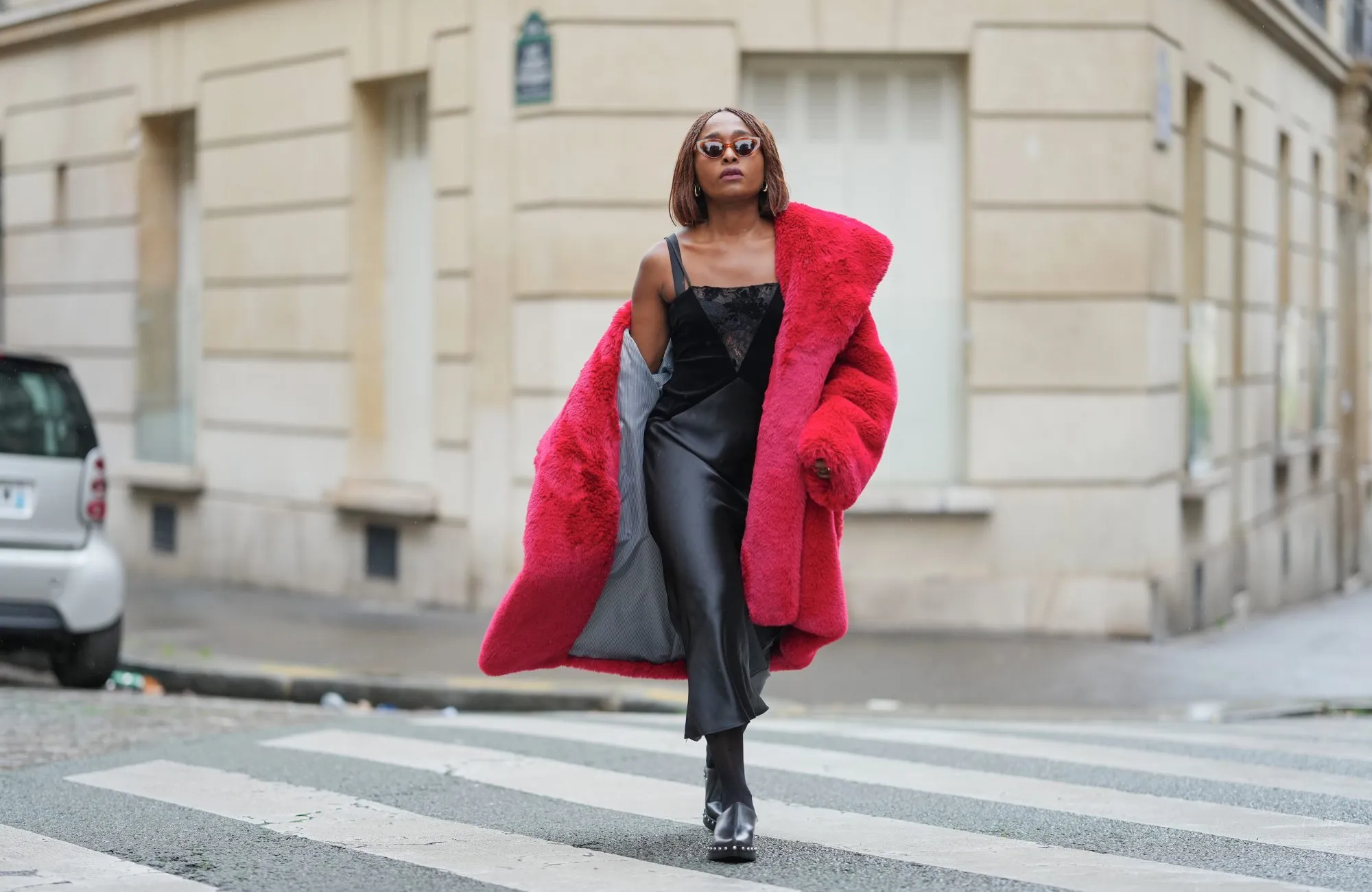 PARIS, FRANCE - DECEMBER 18: Carrole Sagba wears shoulder-length straight brown bob hair with a center part, makeup with an even complexion, defined brows and deep red lipstick, narrow brown-tinted sunglasses, a bright red faux-fur fluffy coat by Milkwhite worn open and draped off the shoulders, a black lace-trim camisole with thin straps, a black leather midi skirt with a straight cut and subtle sheen, sheer black tights, black leather clogs by Zara with a chunky sole, during a street style fashion photo session, on December 18, 2025 in Paris, France. (Photo by Edward Berthelot/Getty Images)