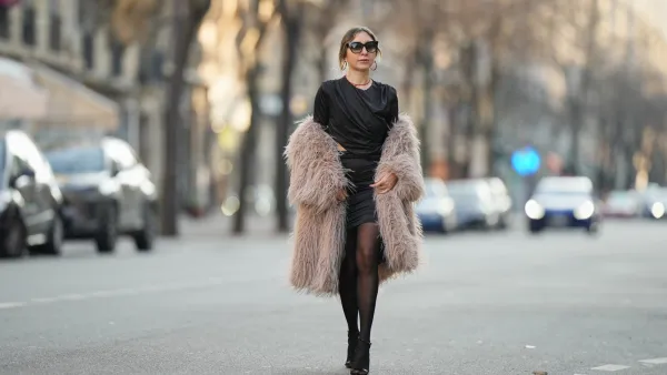 PARIS, FRANCE - DECEMBER 27: Emy Venturini wears black rectangular sunglasses by Dita, earrings, a red necklace, hair pulled back with loose strands framing the face, natural makeup with a soft lipstick finish, a black vintage gathered mini dress with a draped neckline and ruched construction creating an asymmetric silhouette, layered under a taupe beige faux fur coat by Zara worn open, sheer black tights by Calzedonia, and black open-toe ankle shoes with a stiletto heel by Dolce & Gabbana, during a street style fashion photo session, on December 27, 2025 in Paris, France. (Photo by Edward Berthelot/Getty Images)
