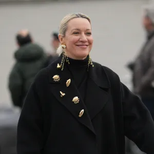 MILAN, ITALY - JANUARY 18: A guest wears hair pulled back into a low bun with a center part and blonde hair with darker roots, long gold dangling chain earrings with shell-like charms and multiple gold metal brooches pinned to the coat lapel, a black woven leather clutch bag, a black turtleneck top layered under an oversized long black wool coat, and black patent leather chunky lug-sole shoes, outside Prada, during Milan Fashion Week - Menswear Fall/Winter 2026-2027, on January 18, 2026 in Milan, Italy (Photo by Edward Berthelot/Getty Images)