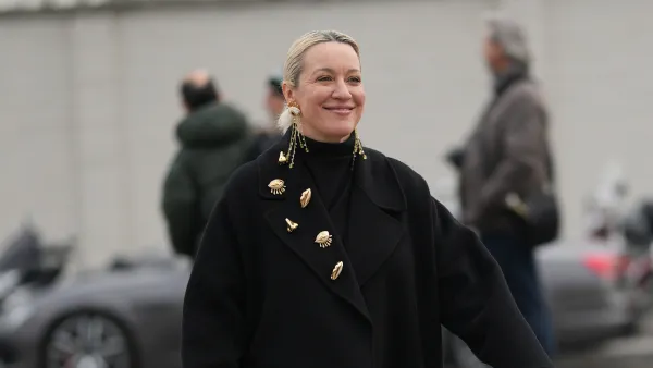MILAN, ITALY - JANUARY 18: A guest wears hair pulled back into a low bun with a center part and blonde hair with darker roots, long gold dangling chain earrings with shell-like charms and multiple gold metal brooches pinned to the coat lapel, a black woven leather clutch bag, a black turtleneck top layered under an oversized long black wool coat, and black patent leather chunky lug-sole shoes, outside Prada, during Milan Fashion Week - Menswear Fall/Winter 2026-2027, on January 18, 2026 in Milan, Italy (Photo by Edward Berthelot/Getty Images)